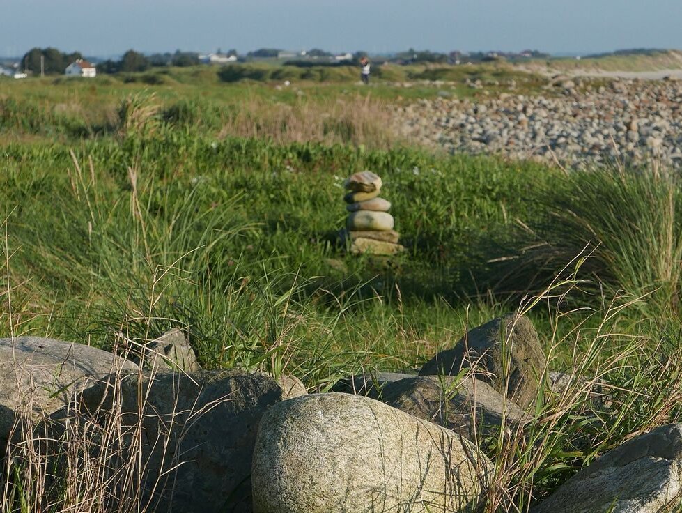 Liten varde ved rullesteinstrand. Grønt gress. Hus i bakgrunnen. 