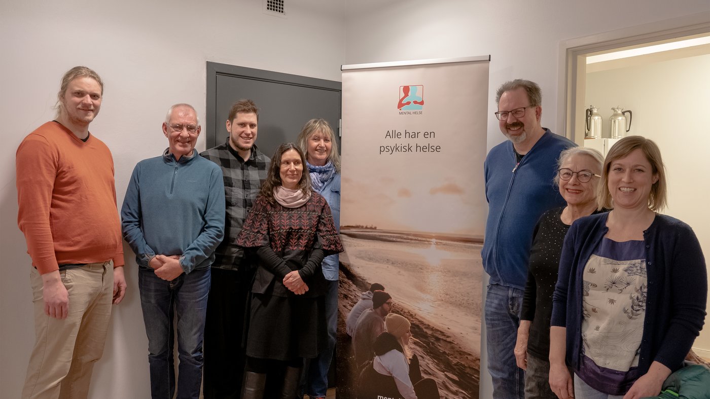 Åtte personer samlet rundt den nye rull-up'n som har teksten "Alle har en psykisk helse" og logoen til Mental Helse. Bakgrunnsfoto er mennesker som ser på en vakker solnedgang i varme farger. 