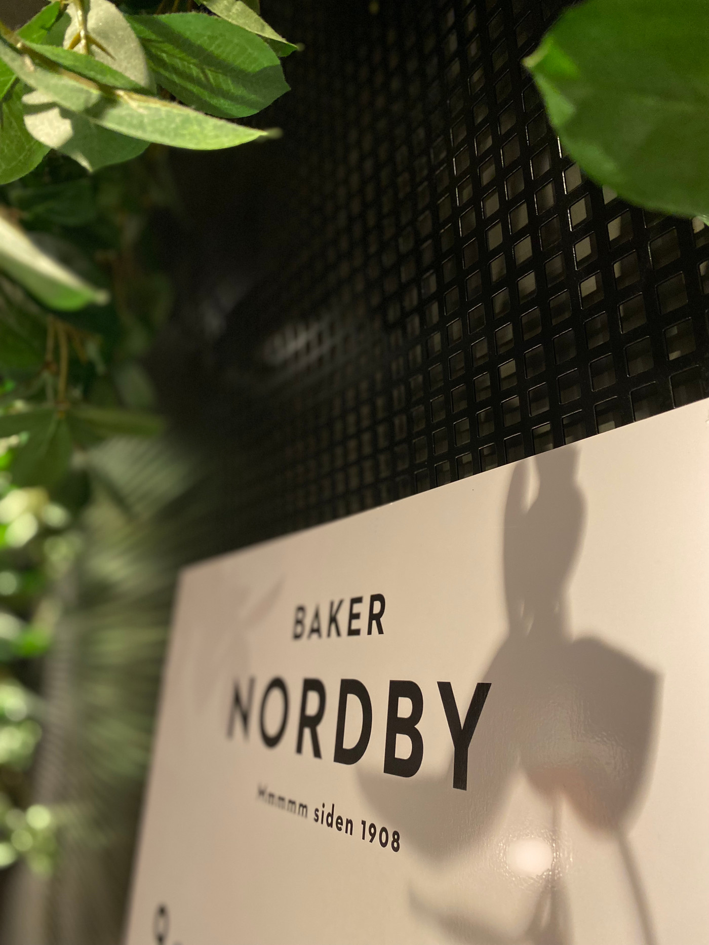 Bilde av et hvitt Baker Nordby-skilt med en grønn plante i bakgrunnen.