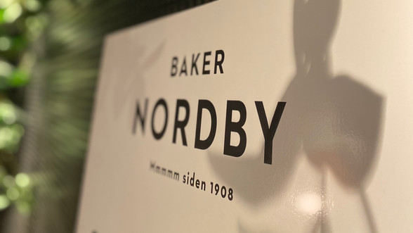 Bilde av et hvitt Baker Nordby-skilt med en grønn plante i bakgrunnen.